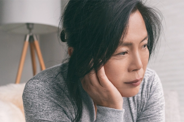 A middle-aged asian woman sitting on her couch, resting her head on her hand, suffering from adrenal fatigue. Get treatment for adrenal fatigue syndrome from ProCare Wellness Institute in Virginia Beach.