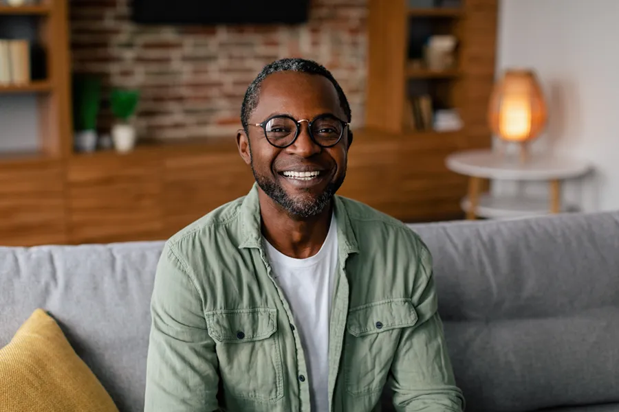 A middle aged black man in a green button up shirt sits on his couch smiling, happy with his treatment for andropause and low testosterone from ProCare Wellness Institute in Virginia Beach.