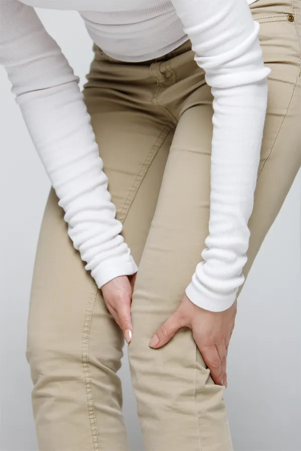 A woman wearing a white long-sleeve shirt and tan pants, clutching her knee, suffering from arthritis. Get arthritis treatment from ProCare Wellness Institute in Virginia Beach.