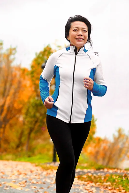 A middle-aged Asian woman jogging in a park during autumn enjoying active lifestyle after successful Hyaluronic Acid Injections for Hip Pain by ProCare Wellness Institute located in Virginia Beach.