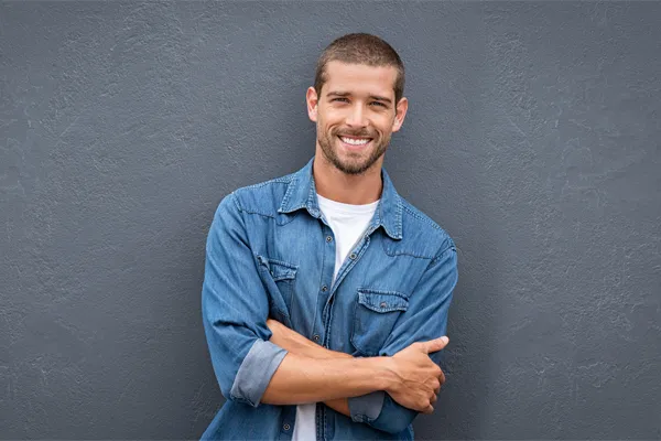 A man in a denim shirt stands smiling against a gray-blue wall, pleased with his testosterone hormone therapy from ProCare Wellness Institute in Virginia Beach.