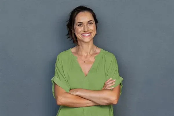 A woman in a green shirt stands smiling against a gray-blue wall, pleased with her hormone therapy from ProCare Wellness Institute in Virginia Beach.
