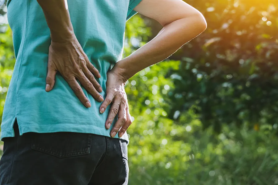A person outdoors, wearing a light turquoise t-shirt, gripping their lower back in a painful gesture. Get Hyaluronic Acid Injections for Back Pain by ProCare Wellness Institute in Virginia Beach.