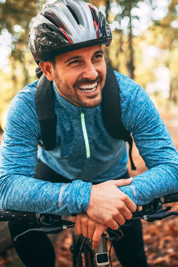 A joyful man on a mountain bike in a forest setting, wearing a helmet and a blue long-sleeved athletic shirt. He is leaning forward on his bike, hands on the handlebars, and wearing a backpack, clearly enjoying his ride after successful Hyaluronic Acid Injections for Back Pain by ProCare Wellness Institute in Virginia Beach.
