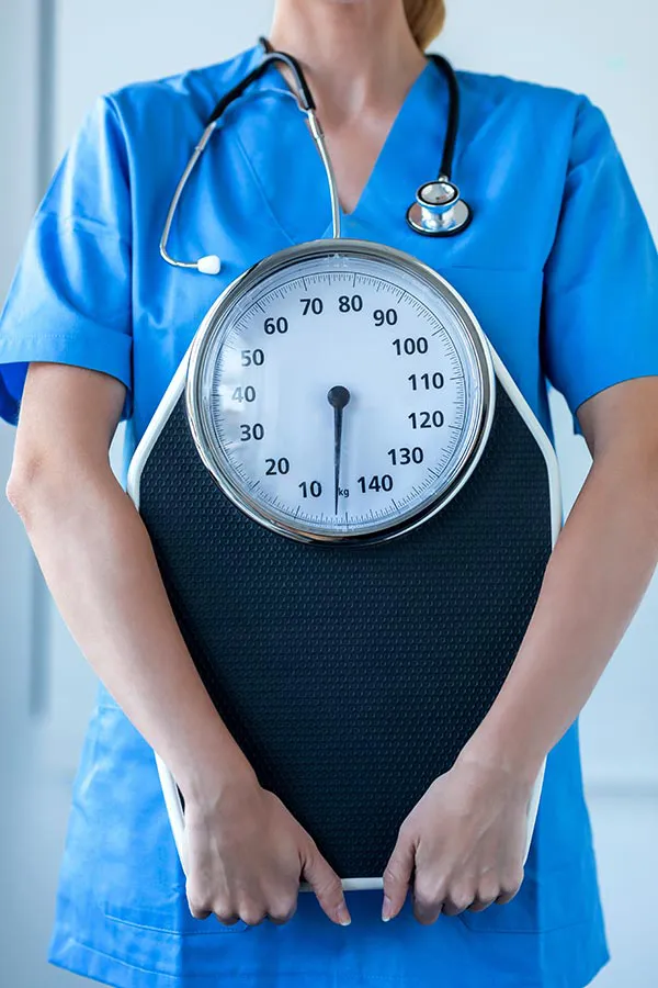 A nurse in blue scrubs with a stethoscope hanging around her neck holds a scale, representing weight loss treatments from ProCare Wellness Institute in Virginia Beach.
