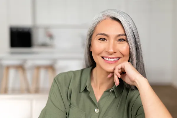 An asian woman with long gray hair and a green shirt sitting and smiling on her couch, happy with her menopause care from [NAME} in Virginia Beach.