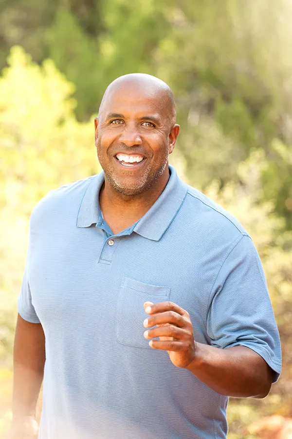 Middle-aged African American man smiling broadly, dressed in a casual blue polo shirt. He is outdoors walking on a sunlit trail happy after receiving advanced Non-Healing Wound Care from ProCare Wellness Institute in Virginia Beach.