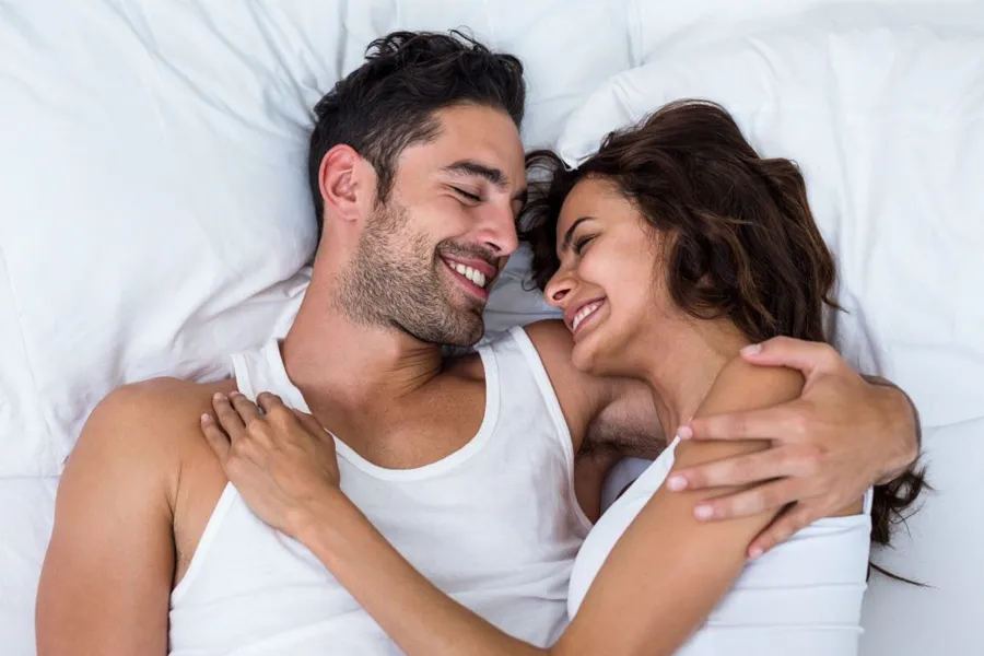 A laughing man and woman in undershirts lie in each other's arms on a white bed, representing successful PT141 sexual wellness therapy for men from ProCare Wellness Institute in Virginia Beach.