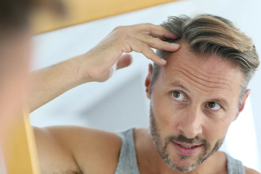 A middle-aged man with blonde and gray hair inspects his receding hairline in the mirror, considering getting peptides for hair restoration treatments from ProCare Wellness Institute in Virginia Beach.