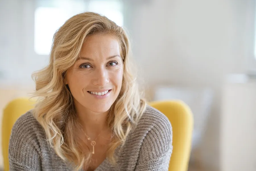 A blonde woman wearing a a gray sweater sits inside in a yellow chair, smiling and happy with her perimenopause treatment from ProCare Wellness Institute in Virginia Beach.