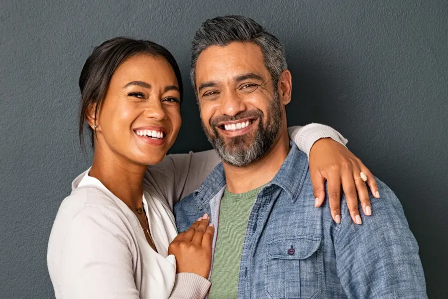 A middle-aged couple with their arms around each other standing against a gray background and smiling, happy with the skin rejuvenation treatments they received from ProCare Wellness Institute in Virginia Beach.