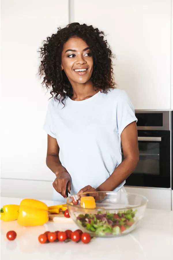 A joyful African American woman preparing food in a modern kitchen. She is chopping yellow bell peppers and has a variety of fresh vegetables like tomatoes and lettuce on the counter, assembling a salad after receiving advanced Non-Healing Wound Treatment by ProCare Wellness Institute in Virginia Beach.