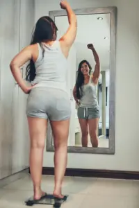 A woman in workout clothes stands on a scale looking in the mirror and pumping her hands in celebration of her successful Tirzepatide treatment from ProCare Wellness Institute in Virginia Beach.